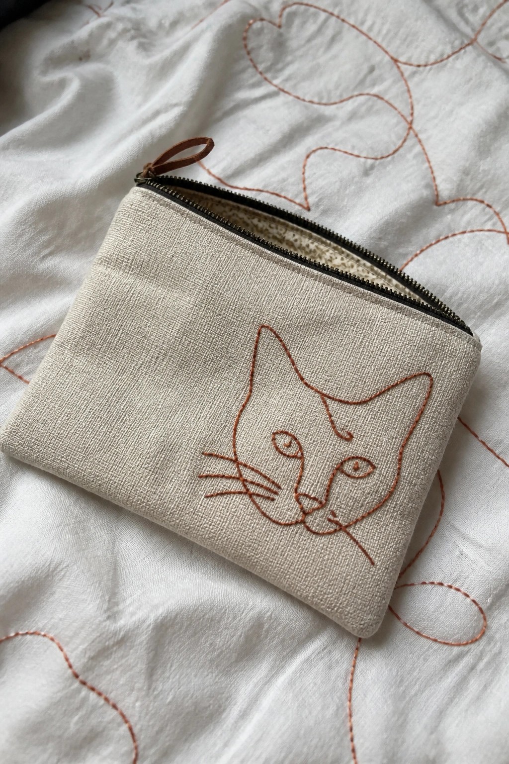 Beige zippered pouch with brown embroidered cat face on white quilted fabric.