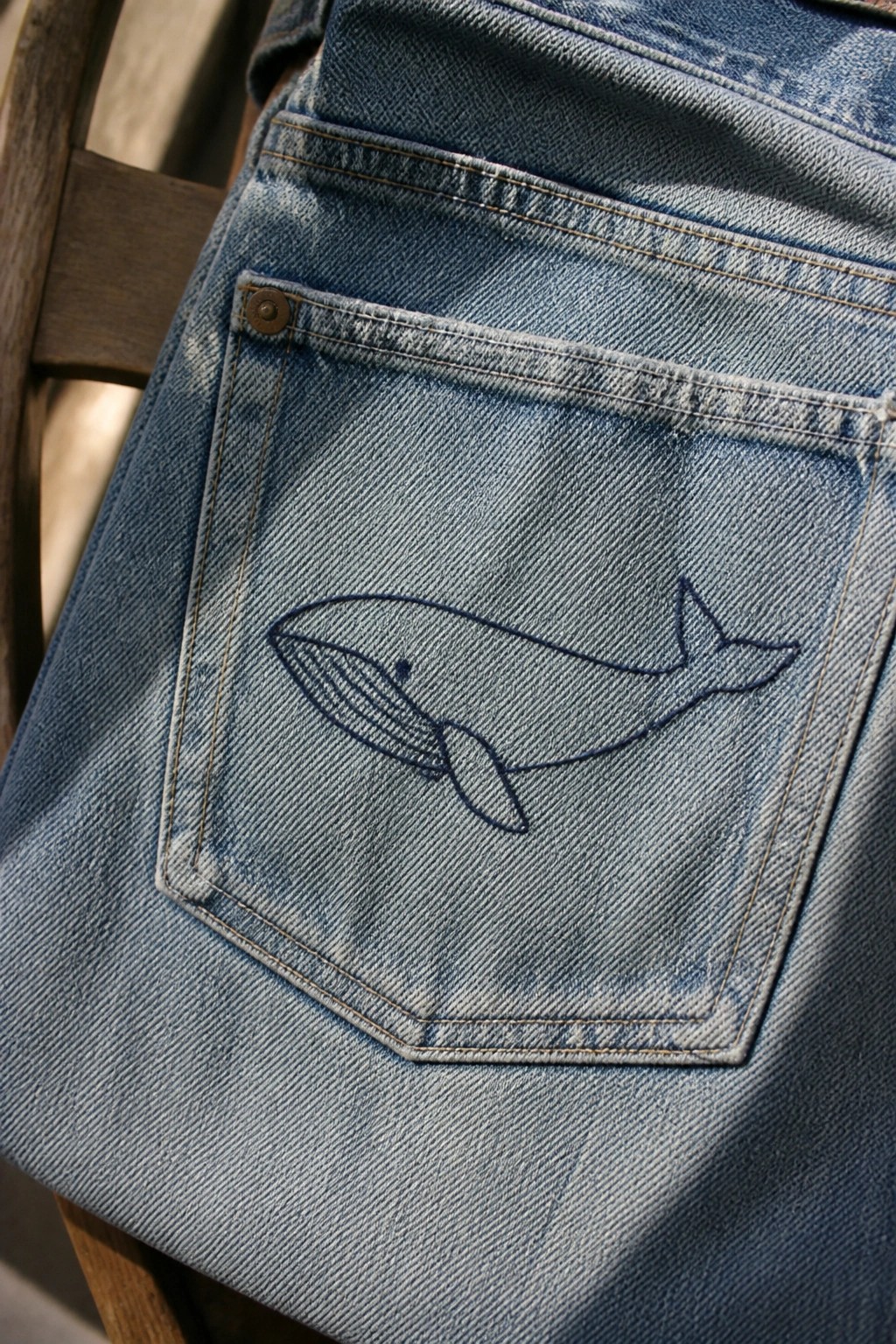 Close-up of blue denim jeans with a dark blue line-art whale embroidered on the back pocket.
