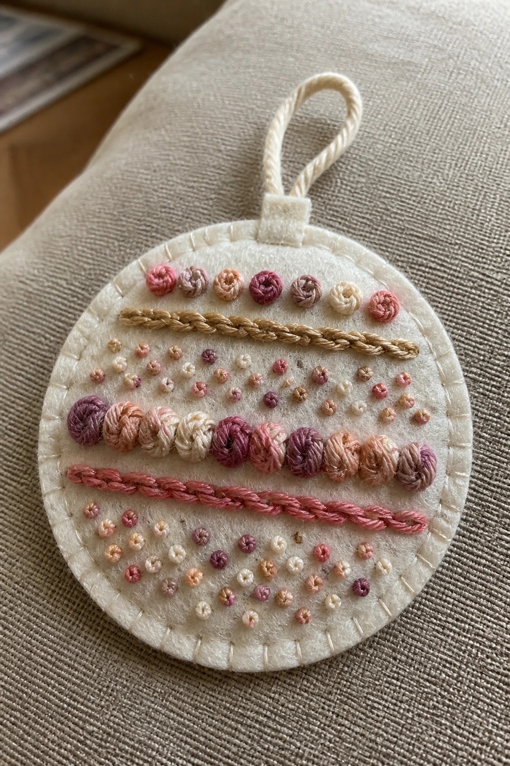 Round white felt ornament with horizontal rows of pink, purple, and beige embroidery stitches and a fabric hanging loop at the top.