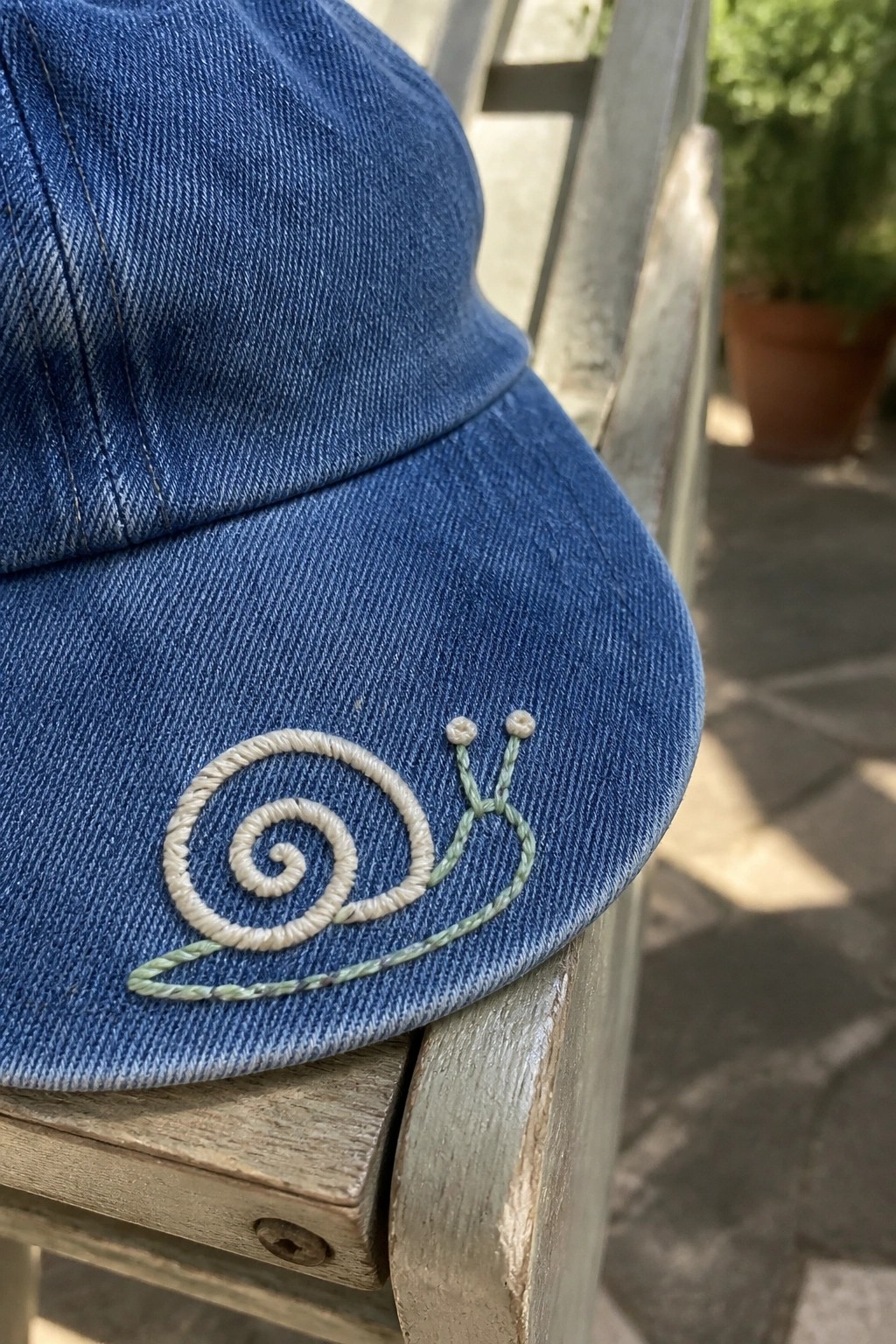 A blue denim cap with a white spiral snail shell and green body embroidered on the brim.