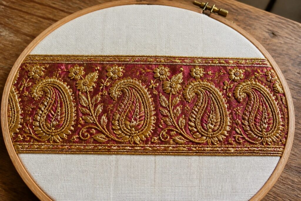 23 Ornate Saree Border Embroidery Designs for Handmade Formalwear Gold paisley embroidery on a magenta band inside a wooden hoop on light fabric.