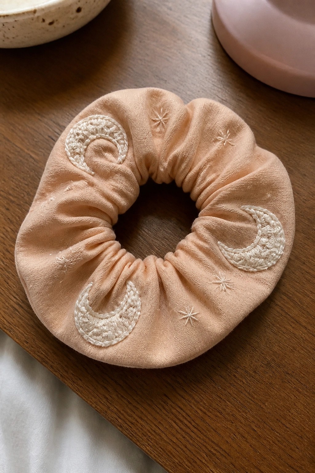 A peach fabric scrunchie with white embroidered crescent moons and small stars lies on a wooden table.