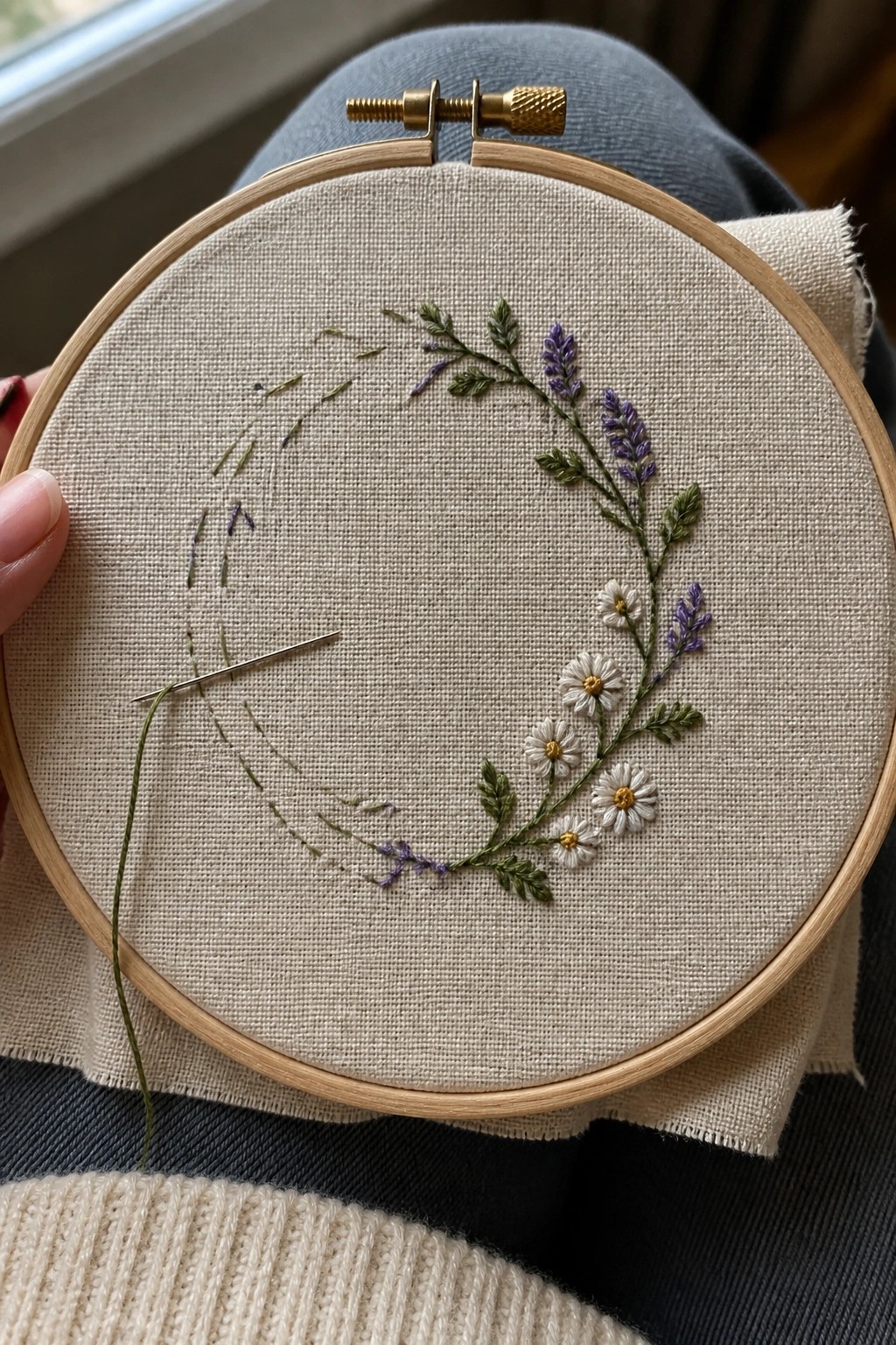 Hands holding wooden hoop stitching lavender daisies on beige fabric over blue jeans and cream sweater