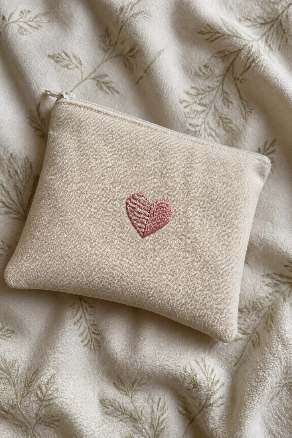 Off-white zippered pouch with pink half-striped heart embroidery on green botanical fabric.