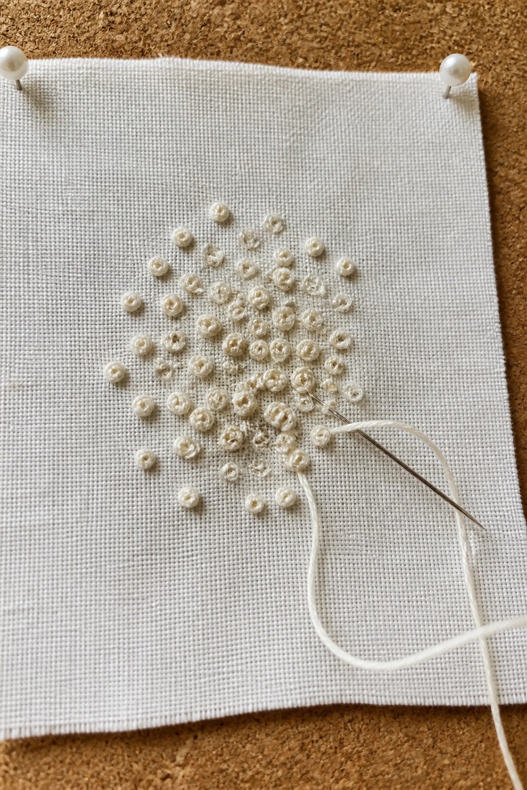 White fabric pinned to cork with pearl pins, white embroidery and needle in progress.
