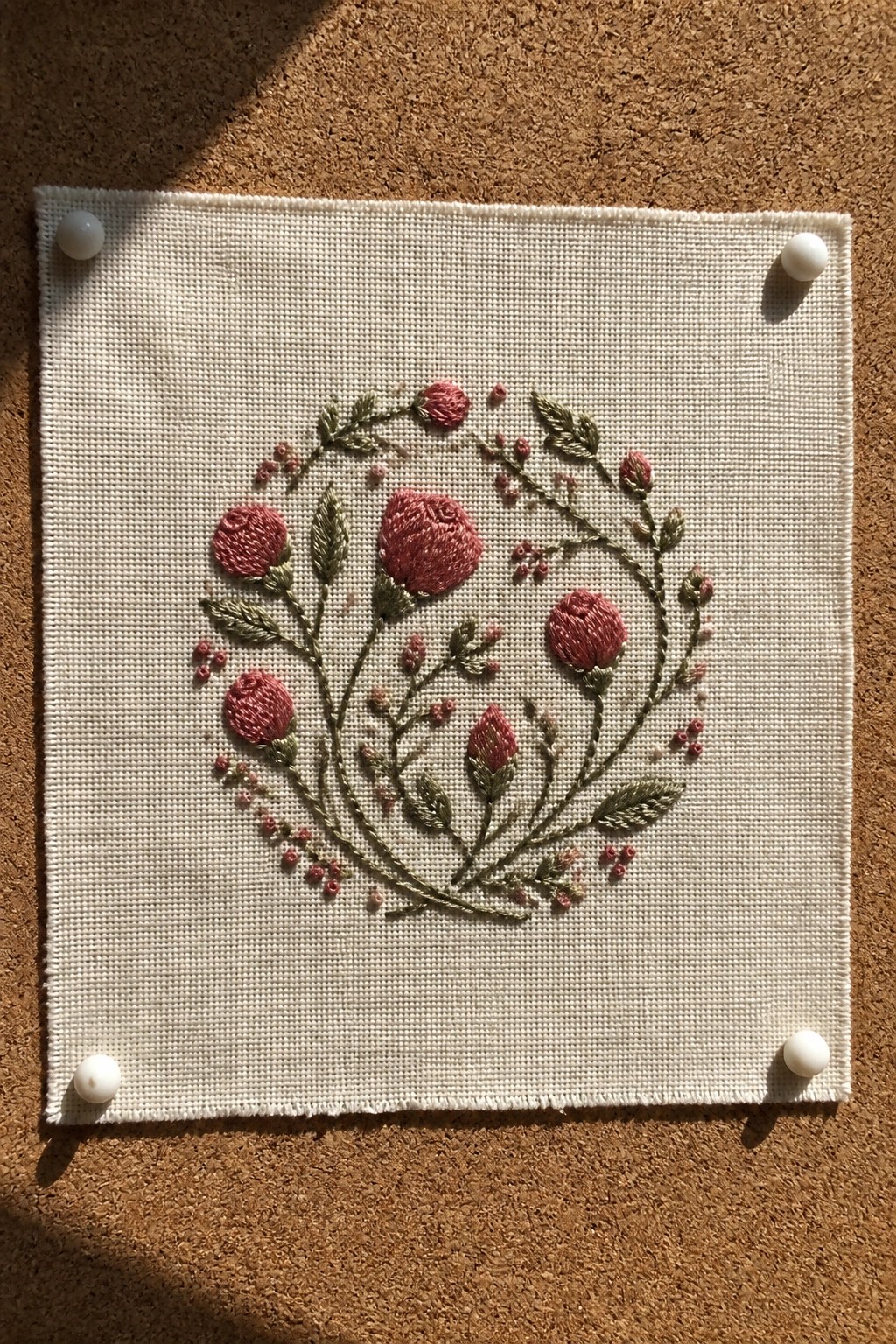 Square of white fabric with a circular pink and green floral embroidery pinned to a corkboard.