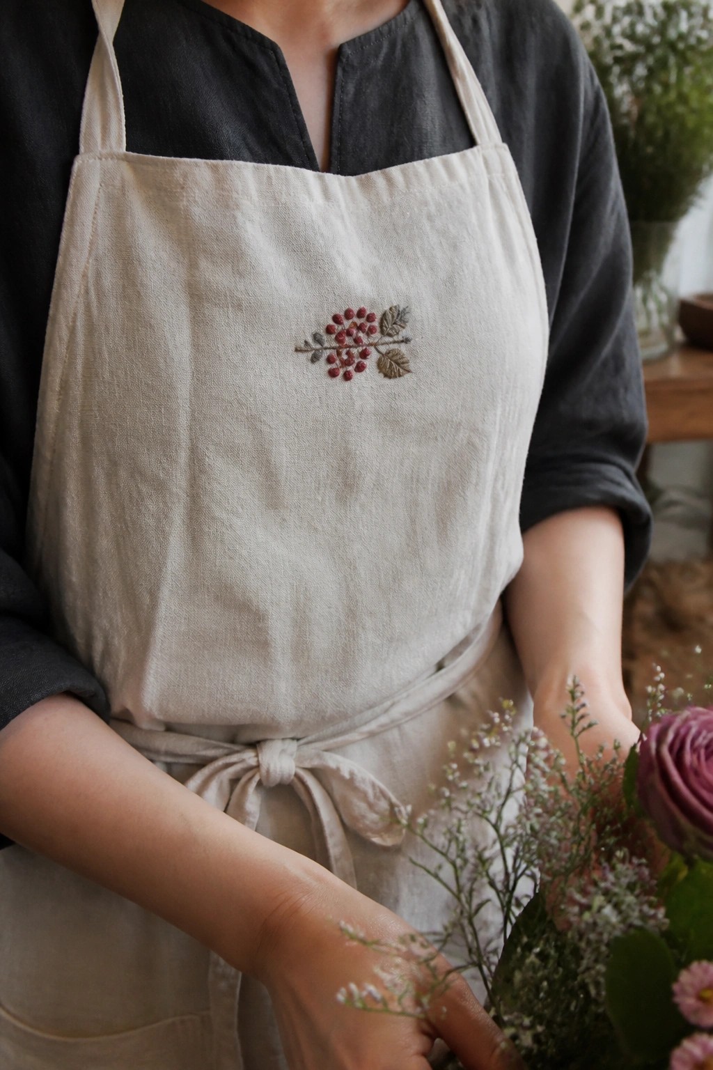 Beige linen apron with a small embroidered branch of red berries and leaves centered on the bib.