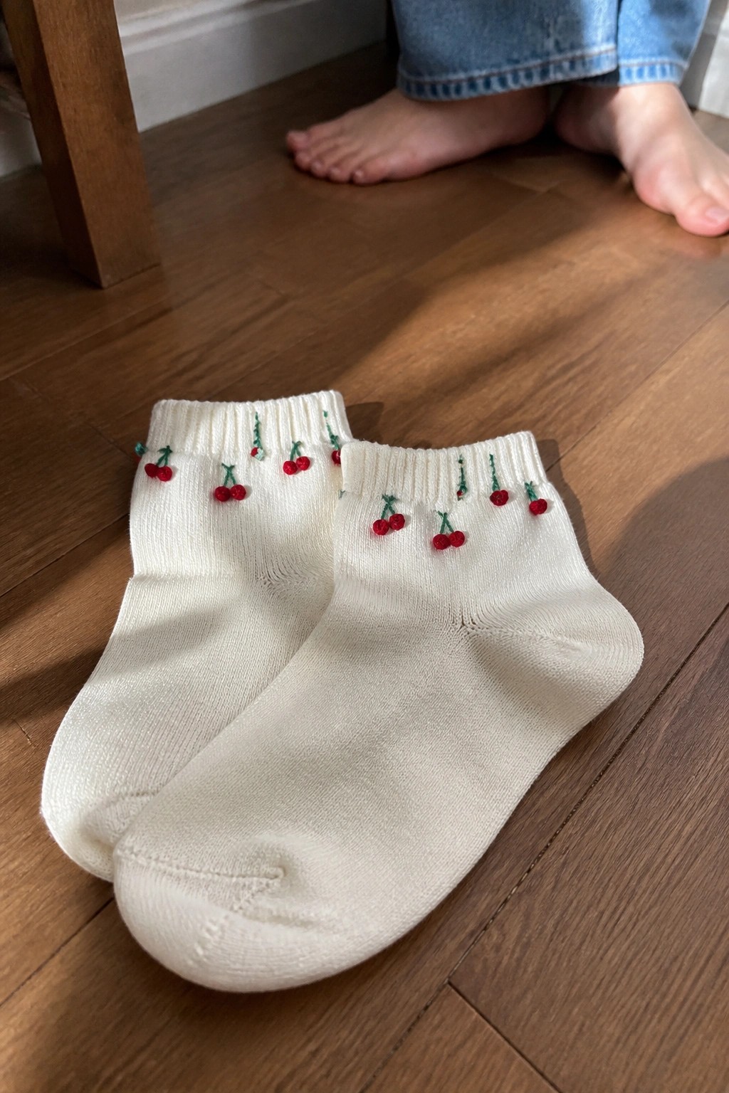 Pair of cream ankle socks with red cherry embroidery on wooden floor near bare feet
