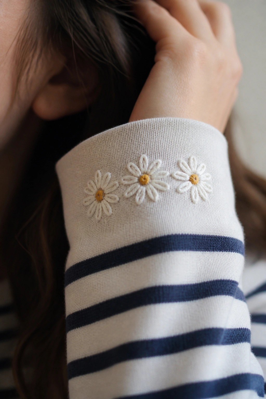 Close-up of a cream sweater sleeve with navy stripes and three small white daisies embroidered on the cuff.