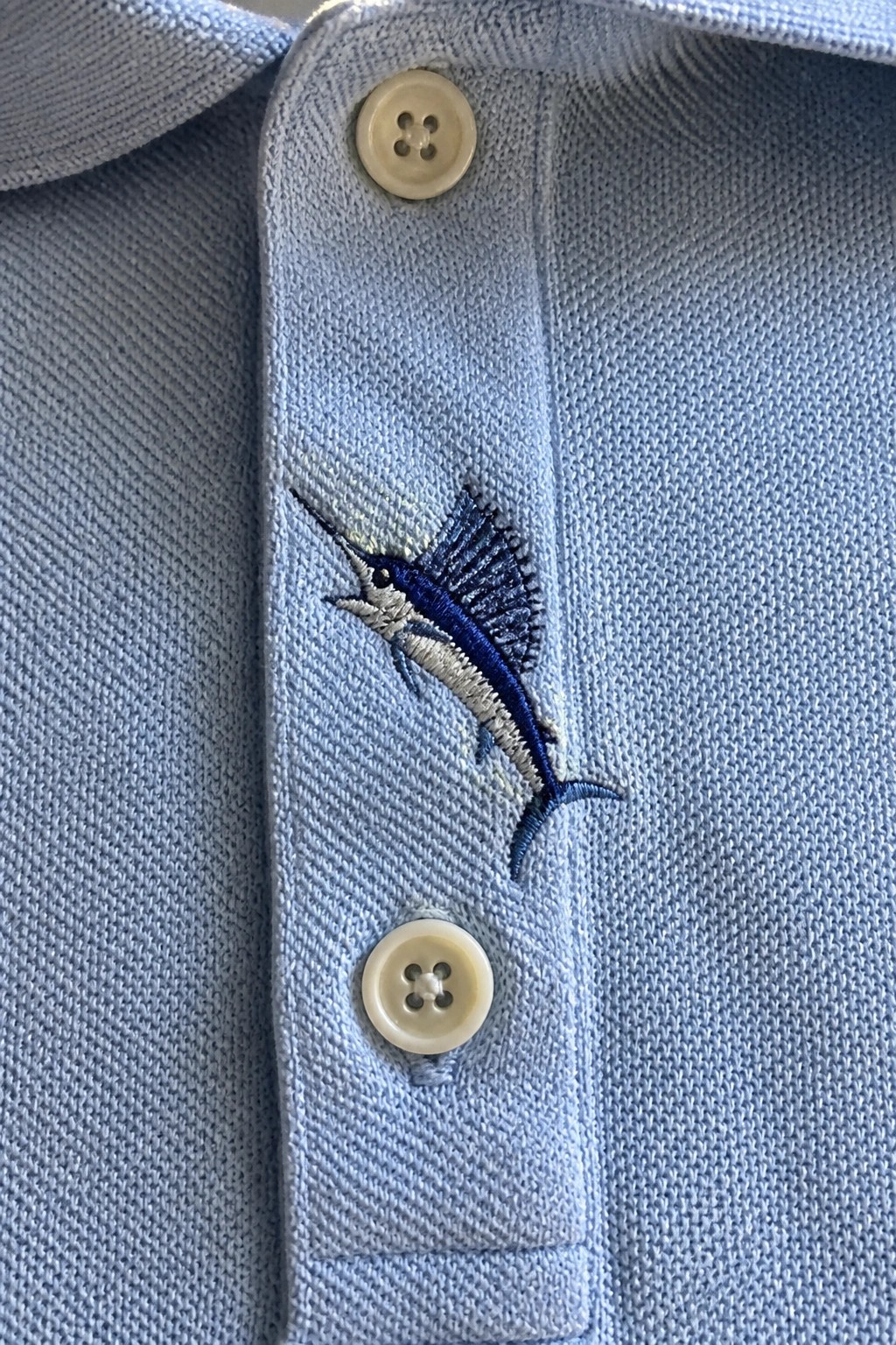 Light blue polo shirt with blue marlin embroidery between white buttons