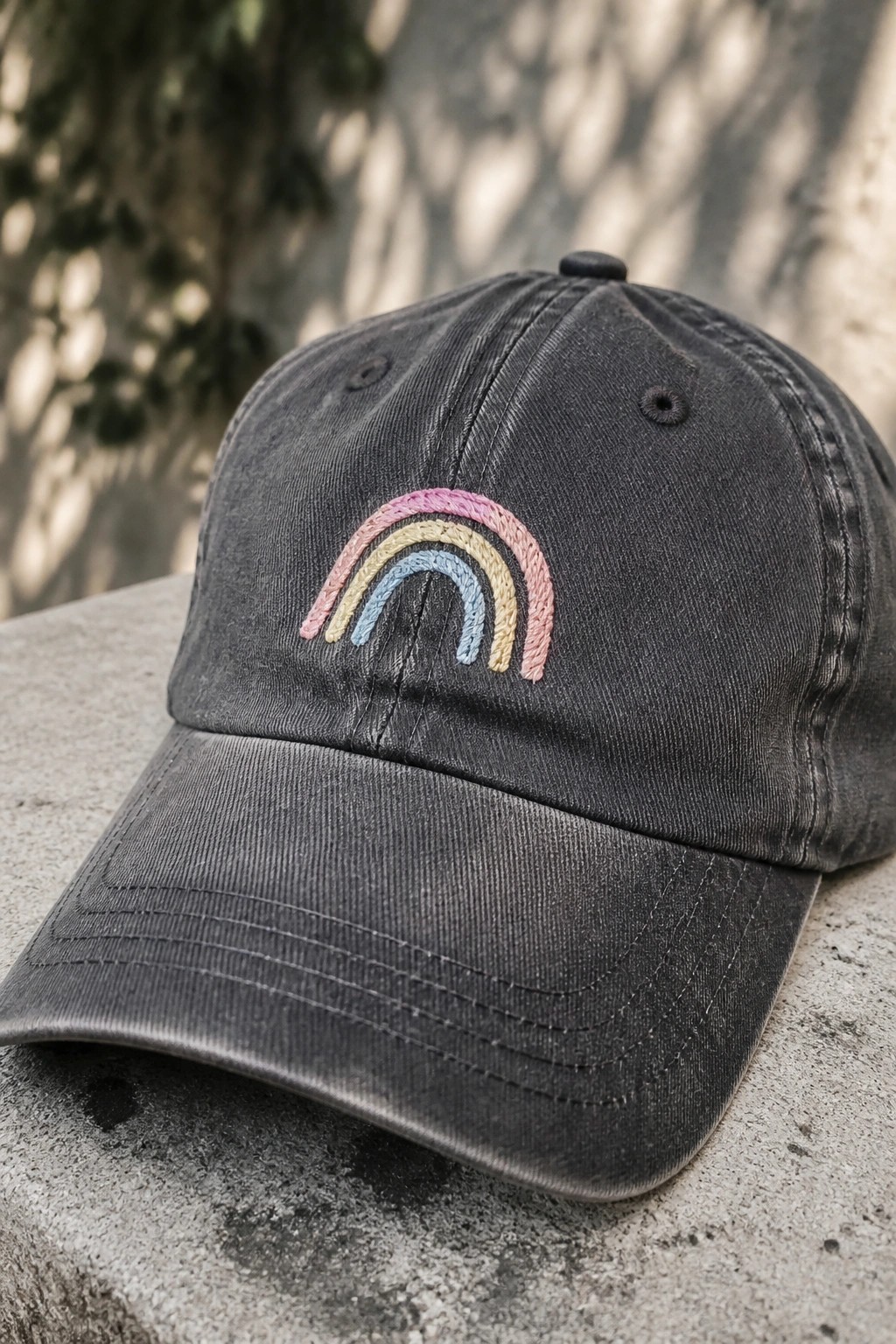 Distressed dark gray baseball cap with pastel rainbow embroidery on stone surface