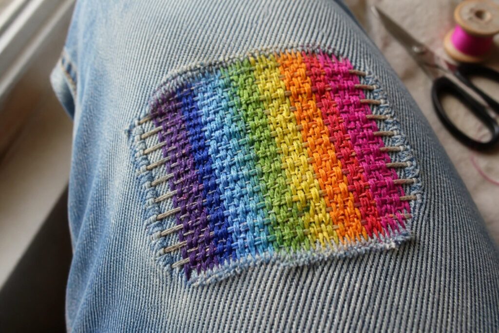 Rainbow embroidered patch repairing tear in blue denim jeans, scissors nearby.