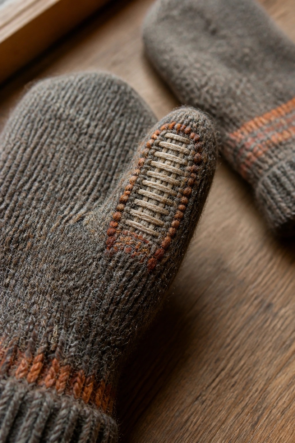 Gray knitted mittens with orange stripes and beige darning on thumb, on wood.