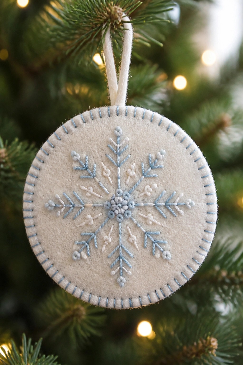 Felt Christmas ornament with light blue embroidered snowflake hanging from pine branch.