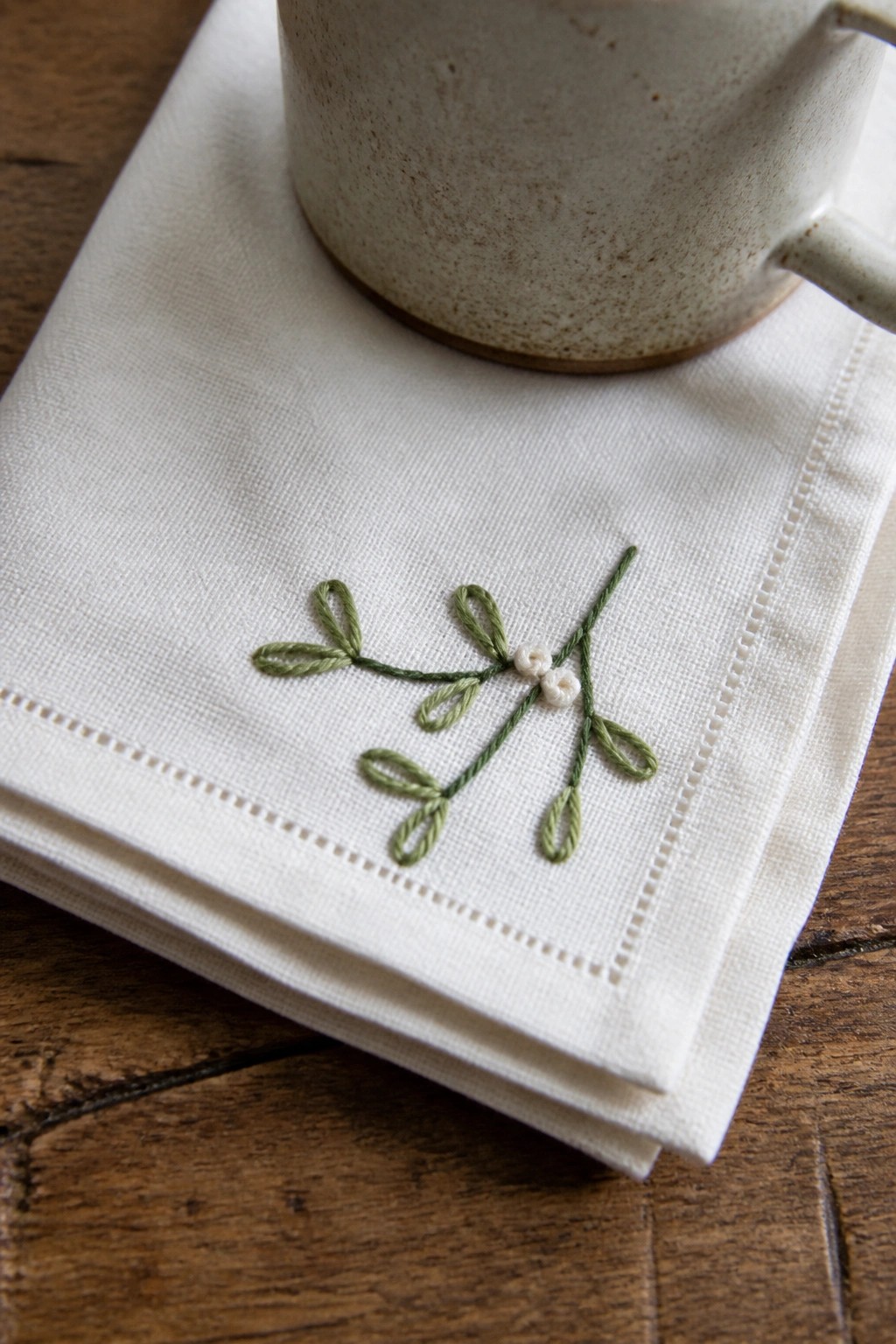 White napkin with green embroidered mistletoe under speckled ceramic mug on wood