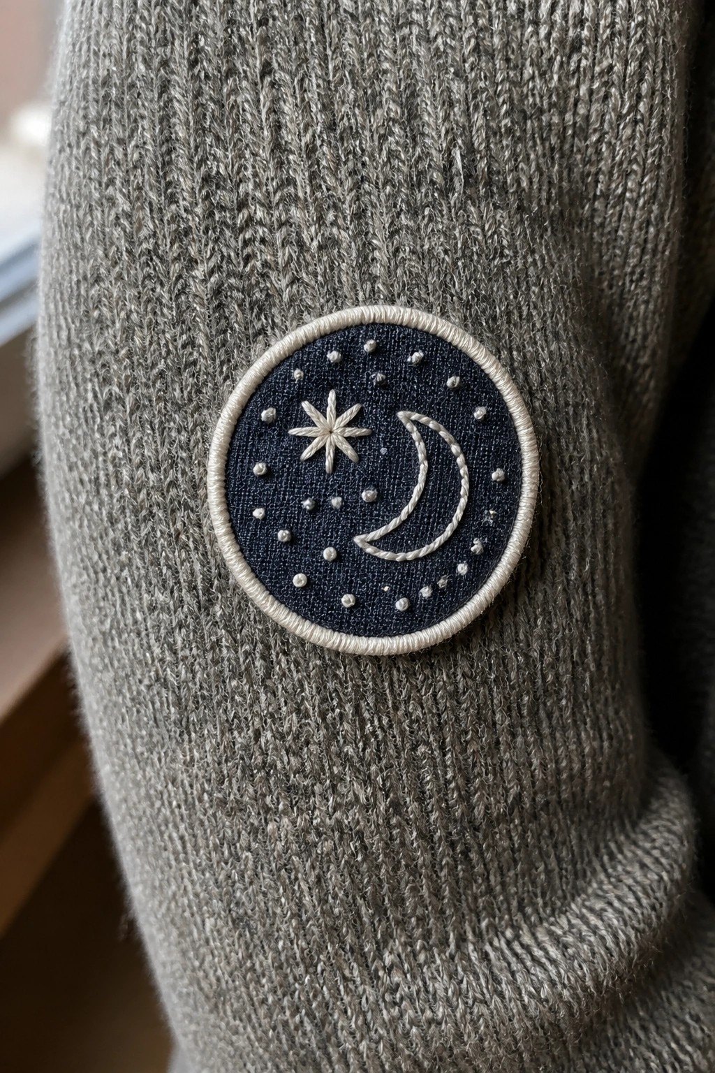 Gray knit sweater sleeve with embroidered navy patch of moon, star, and dots.