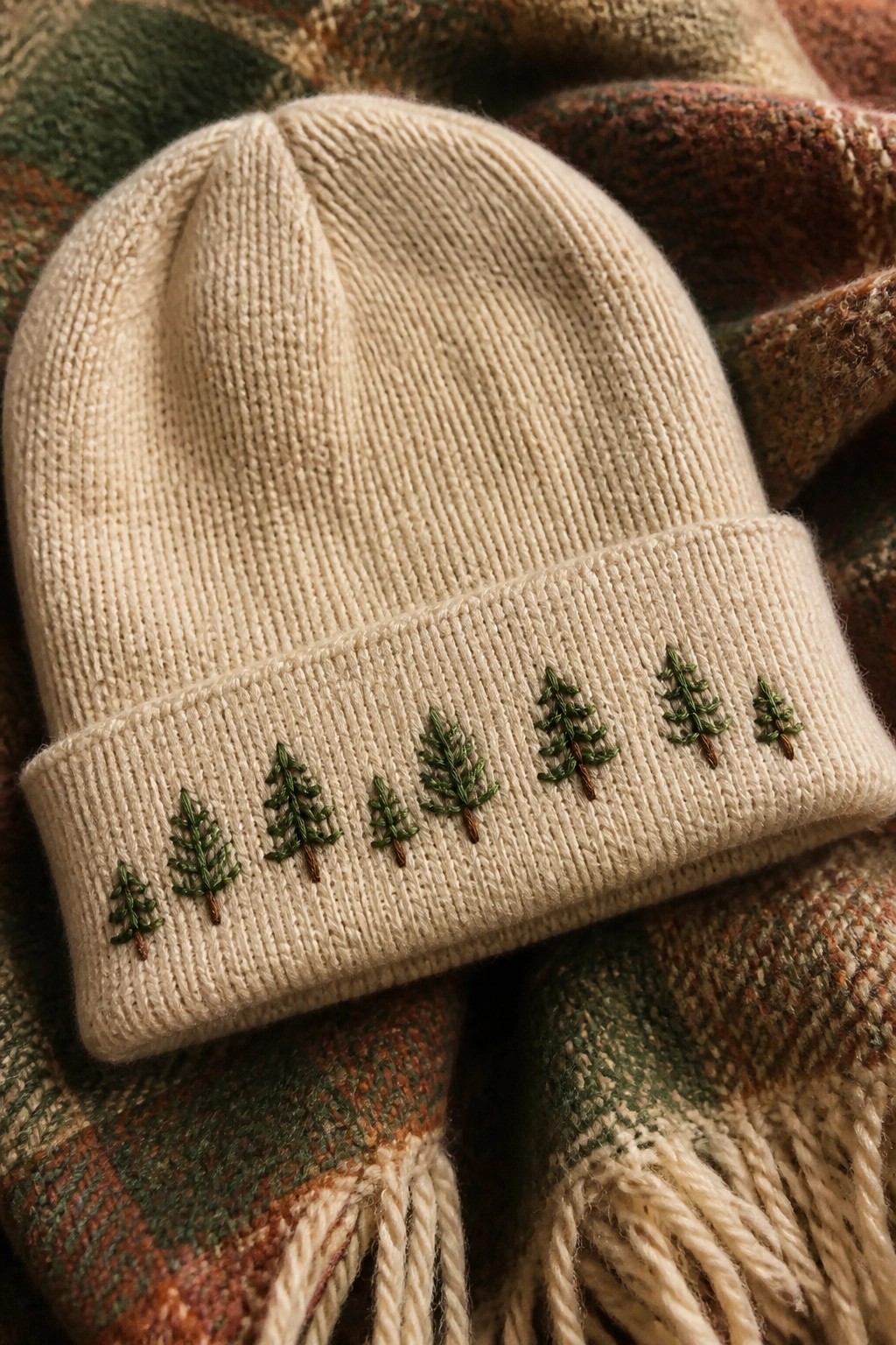 Cream knit beanie with a row of small green embroidered pine trees along the brim on a plaid blanket.
