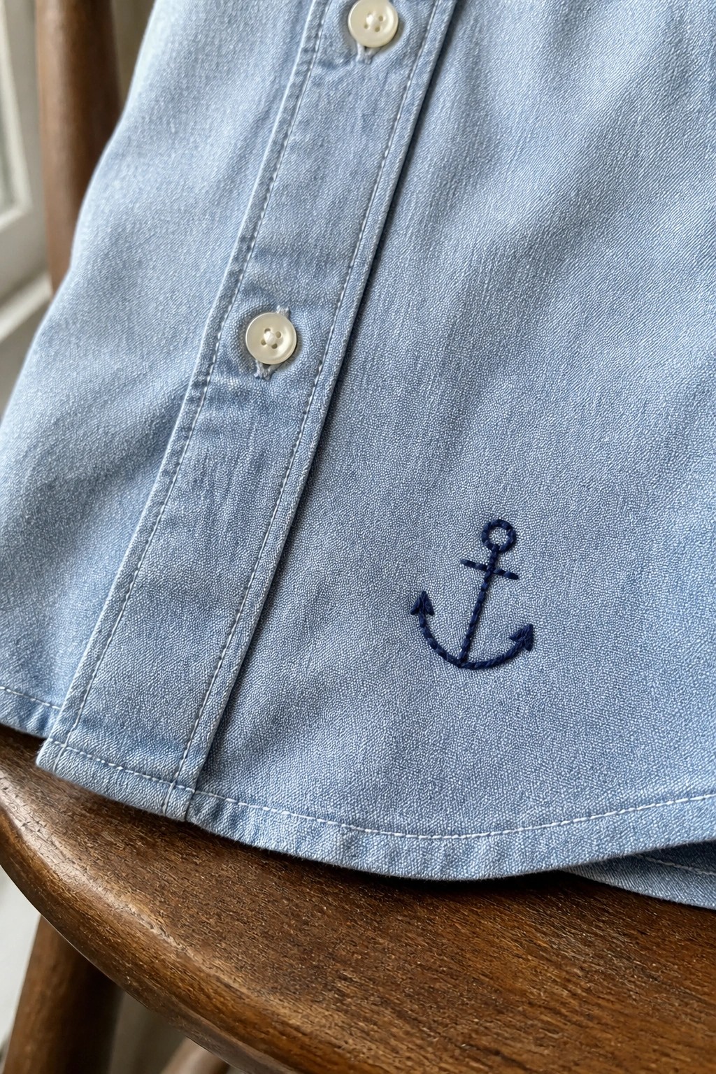 Light blue button-down shirt with navy embroidered anchor and white buttons on wood