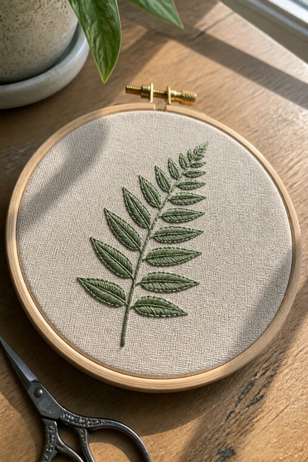 Wooden hoop with green embroidered leaf on beige fabric beside scissors on table.