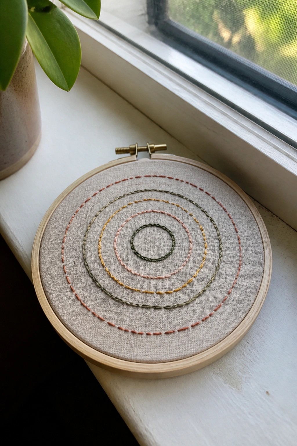 Embroidery hoop with fabric showing concentric circles stitched in multiple muted colors resting on a windowsill.