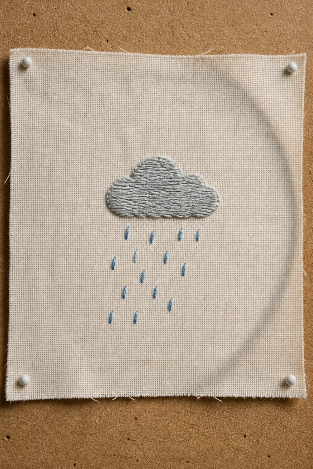 Gray embroidered cloud with blue raindrops on pinned off-white fabric