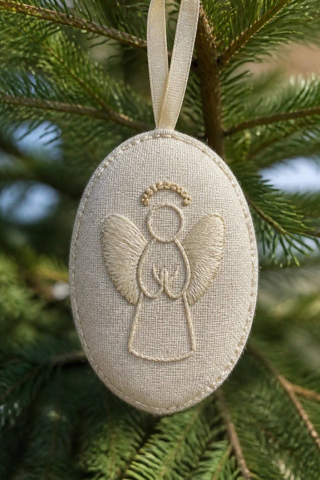Embroidered angel with halo and wings on oval beige fabric ornament on pine branch