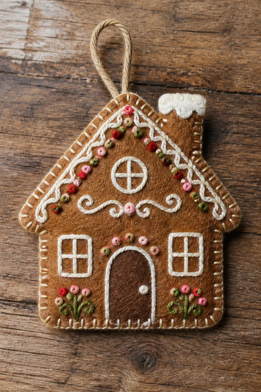 Handmade brown felt gingerbread house ornament with white embroidery and colorful beads on wood.