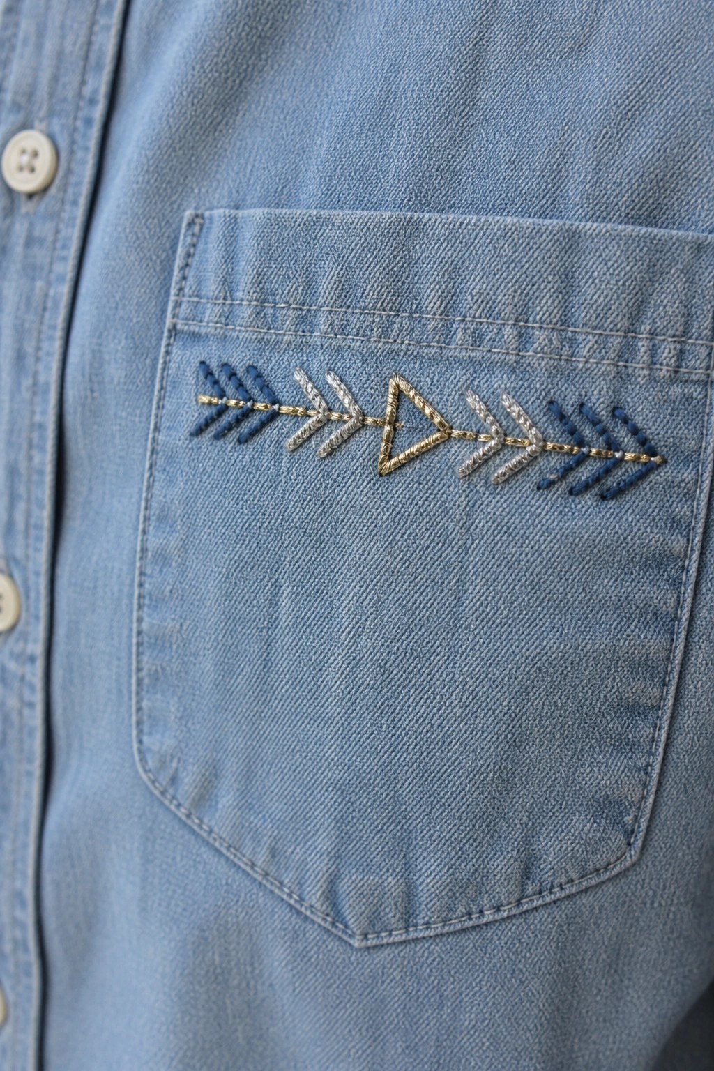 Close-up of blue and gold arrow embroidery running across the top of a light blue denim shirt pocket.