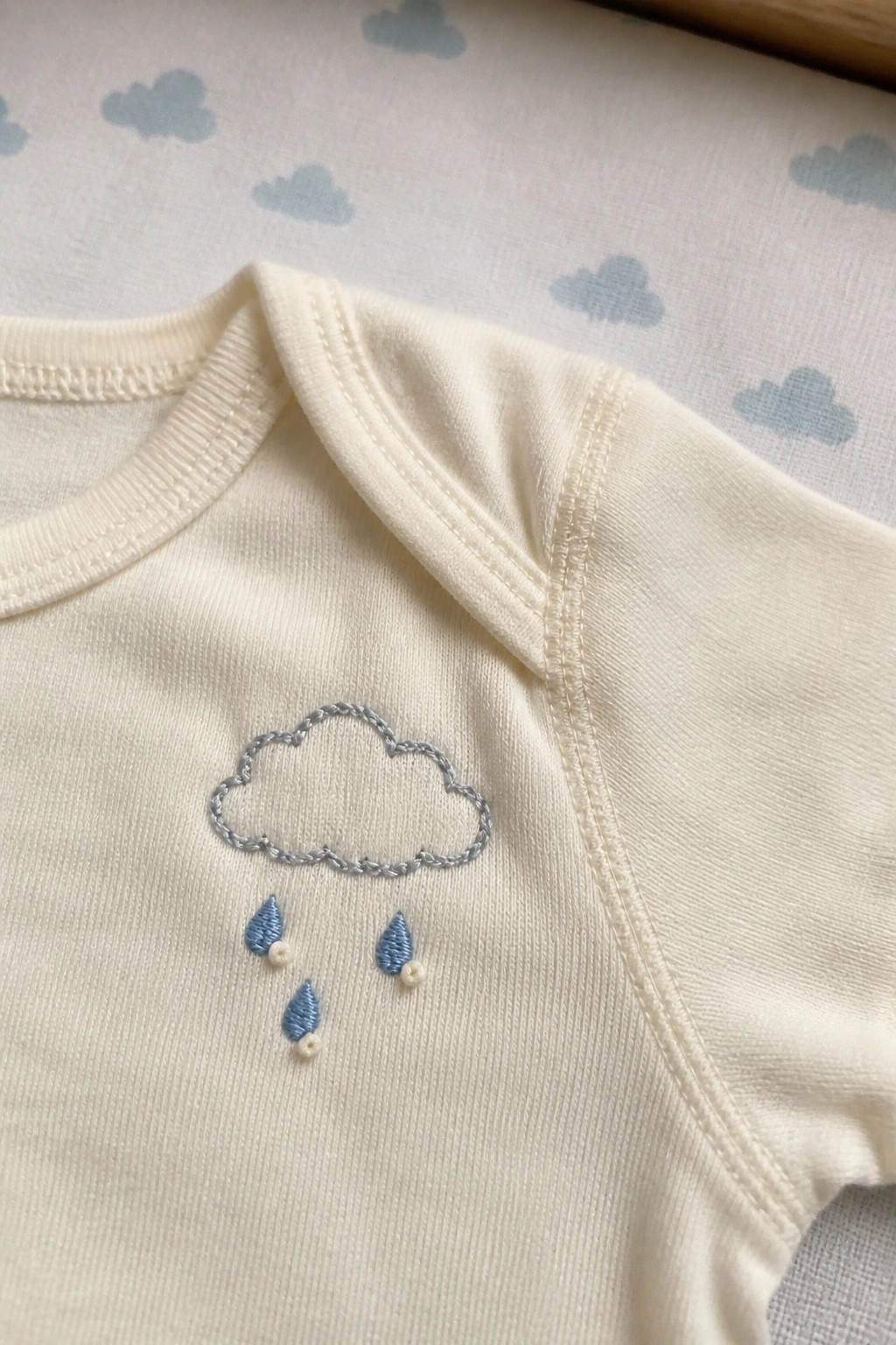 Cream baby onesie with embroidered gray cloud and blue raindrops on cloud-patterned fabric