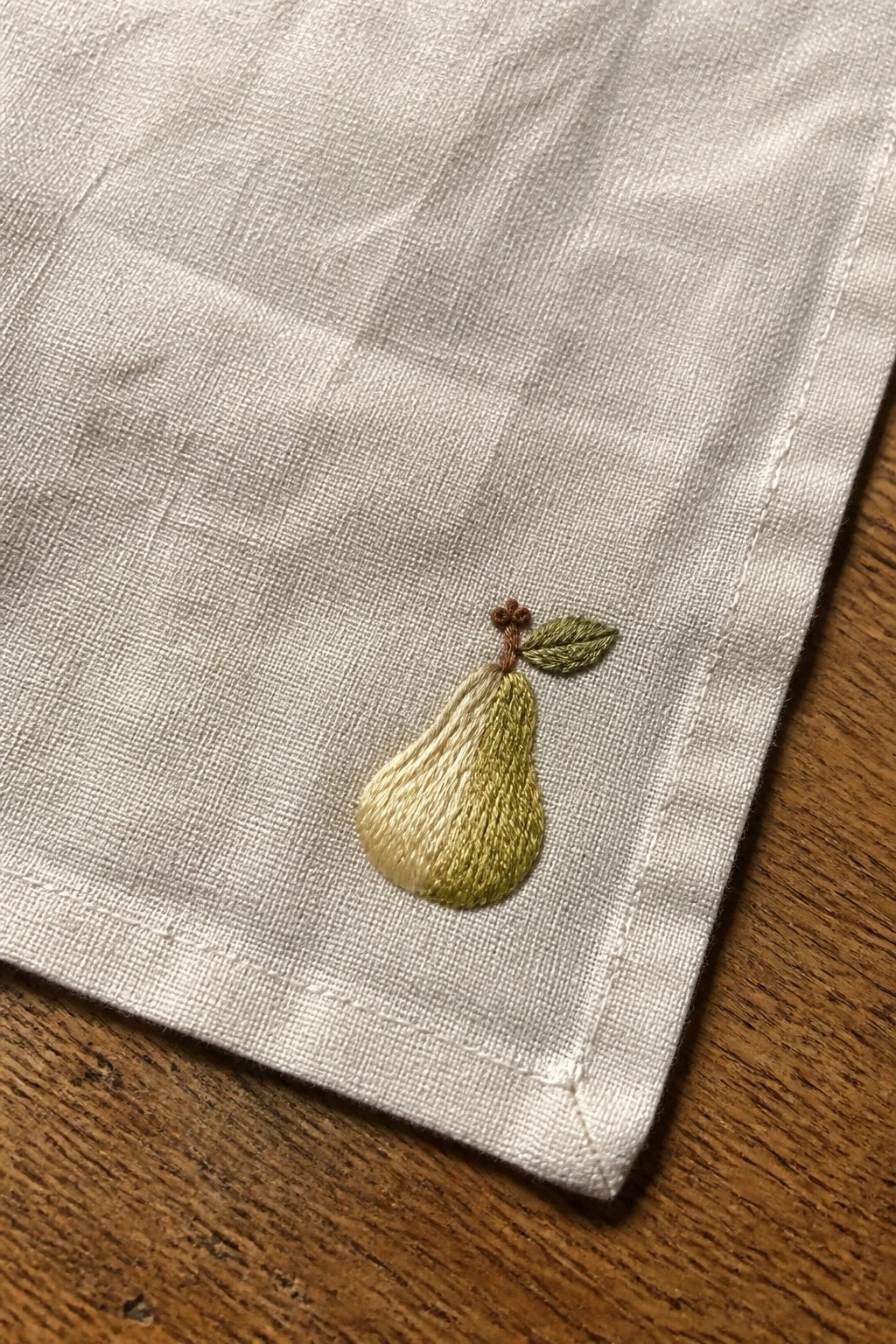 Yellow-green embroidered pear with brown stem on white linen fabric.
