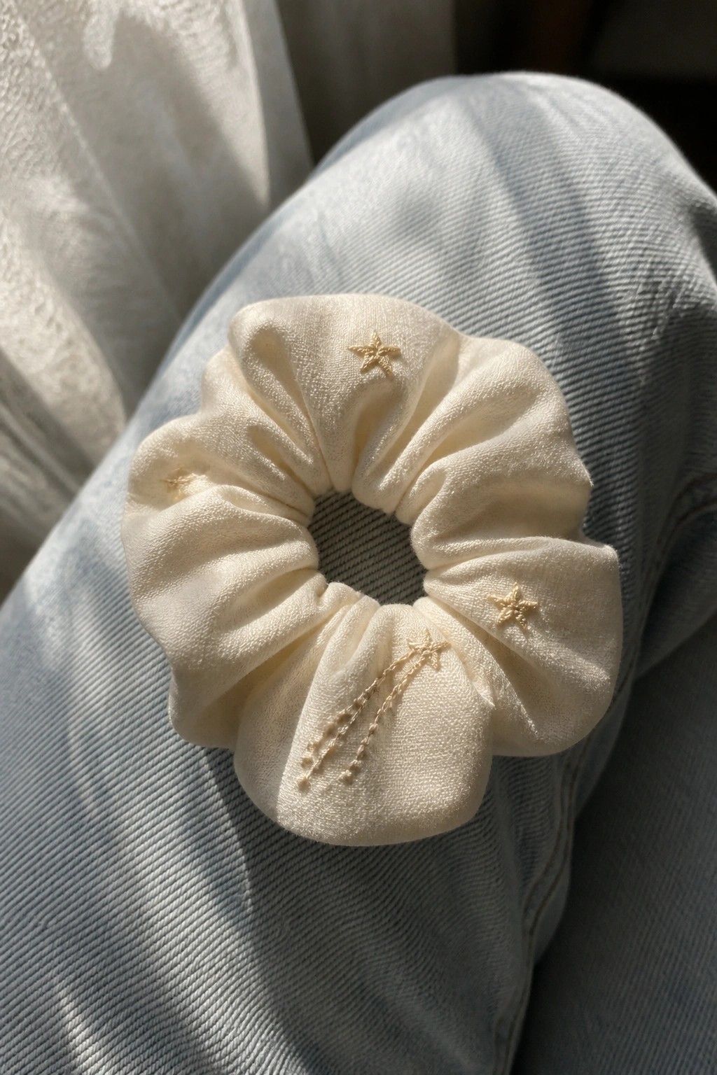 A cream fabric scrunchie with small gold embroidered stars sits on blue denim.