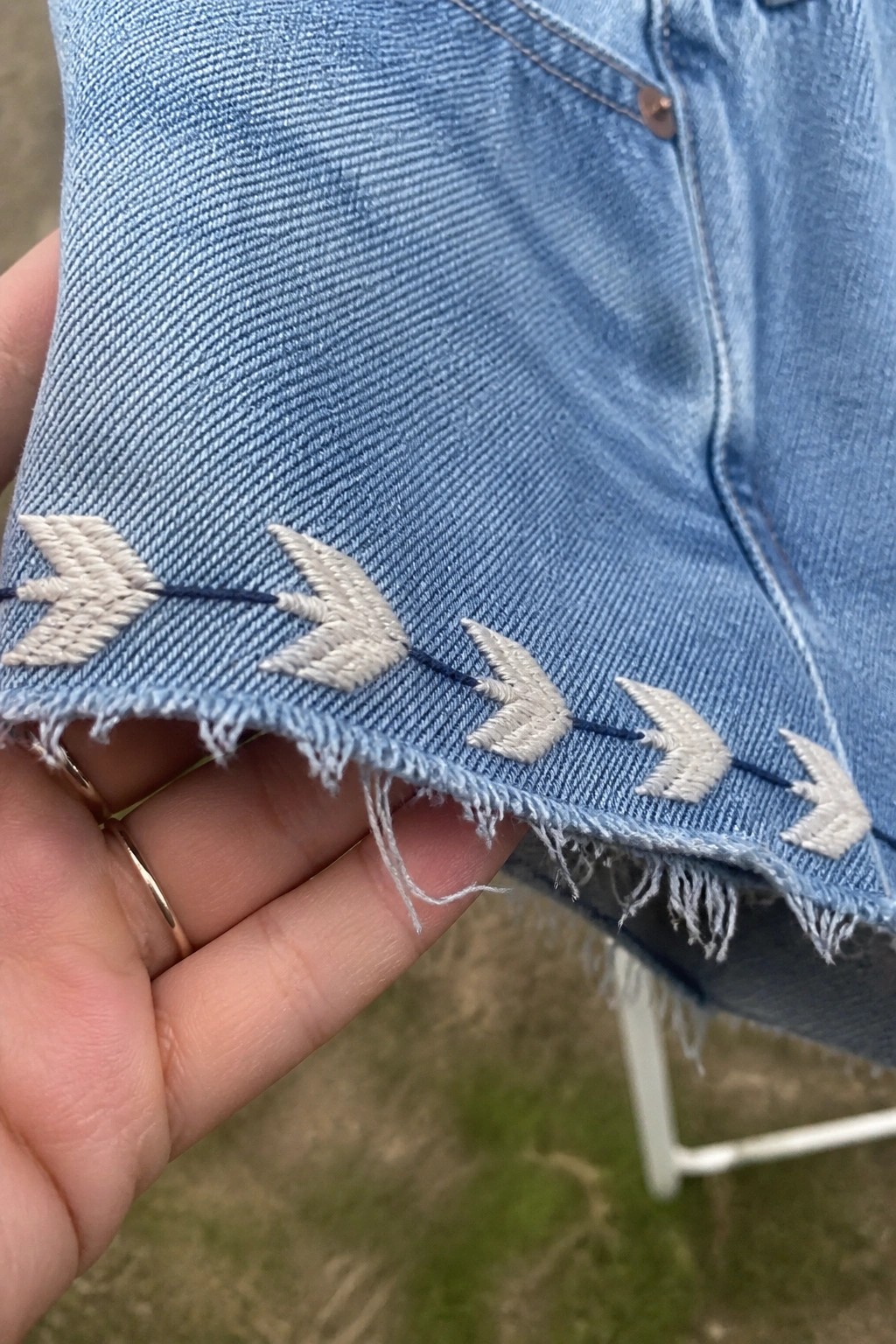Close-up of white arrow shapes embroidered in a line along the frayed hem of light blue denim, held by a hand wearing two gold rings.