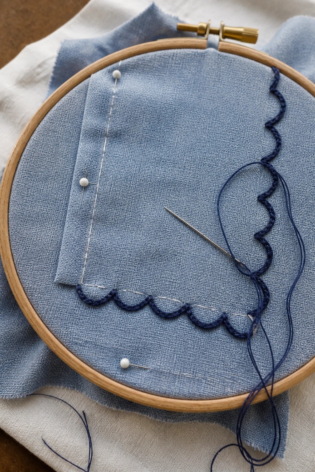 Wooden hoop with light blue fabric, dark blue scalloped stitching, and needle.