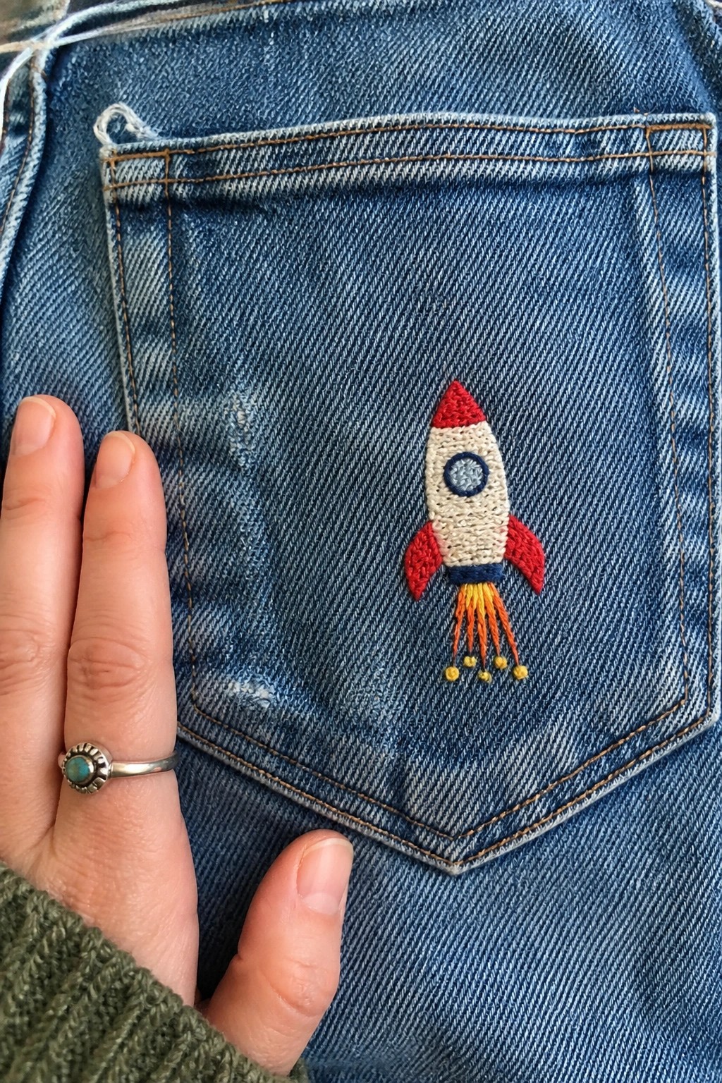 Blue denim jeans pocket with colorful embroidered rocket beside ringed hand