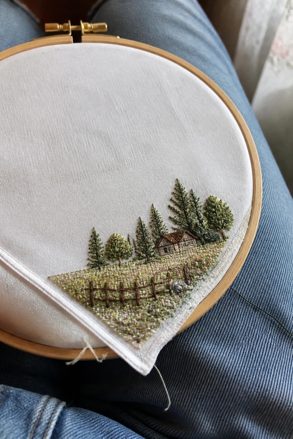 Wooden hoop with embroidered house, trees, and fence on blue denim lap