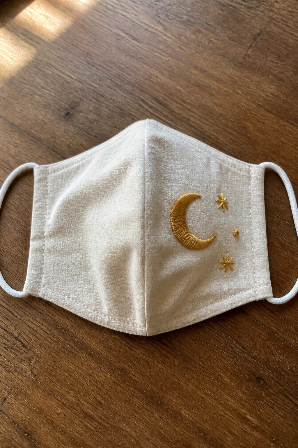 White fabric face mask with gold crescent moon and stars embroidery on wood.