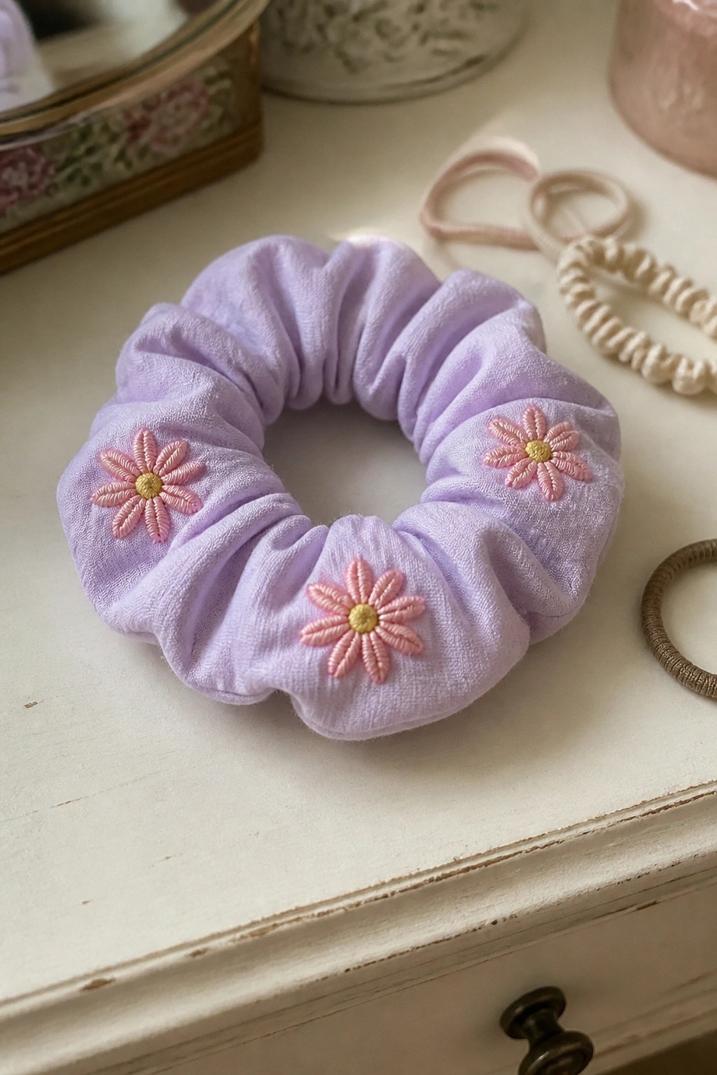 A lavender scrunchie with three small pink and yellow embroidered flowers spaced around the band.