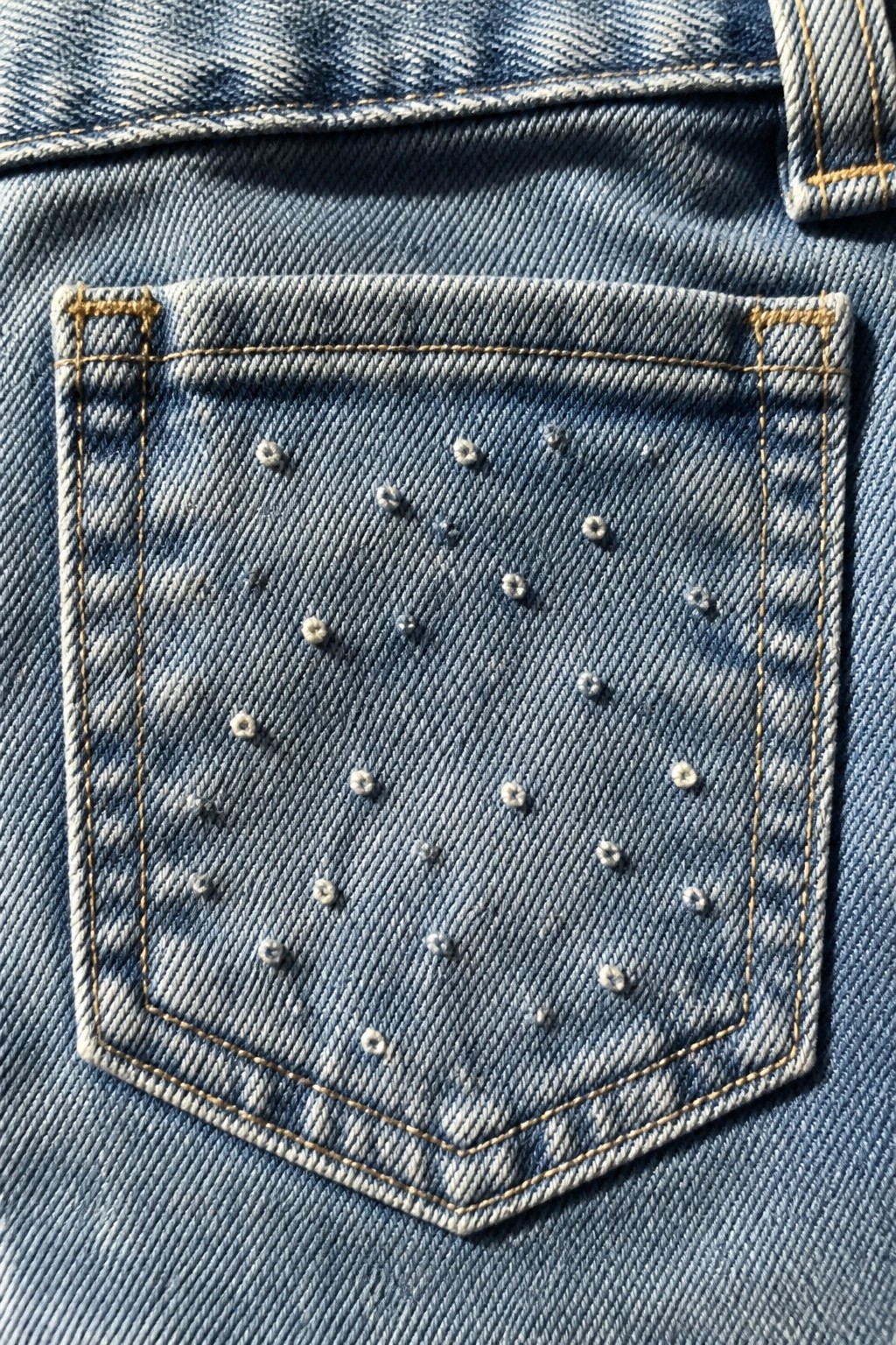 Blue denim jeans pocket with yellow stitching and scattered white studs.