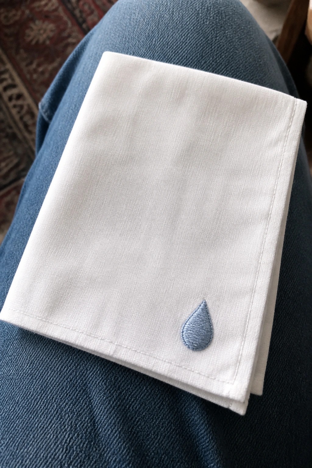 White folded handkerchief with light blue teardrop embroidery on blue denim lap.