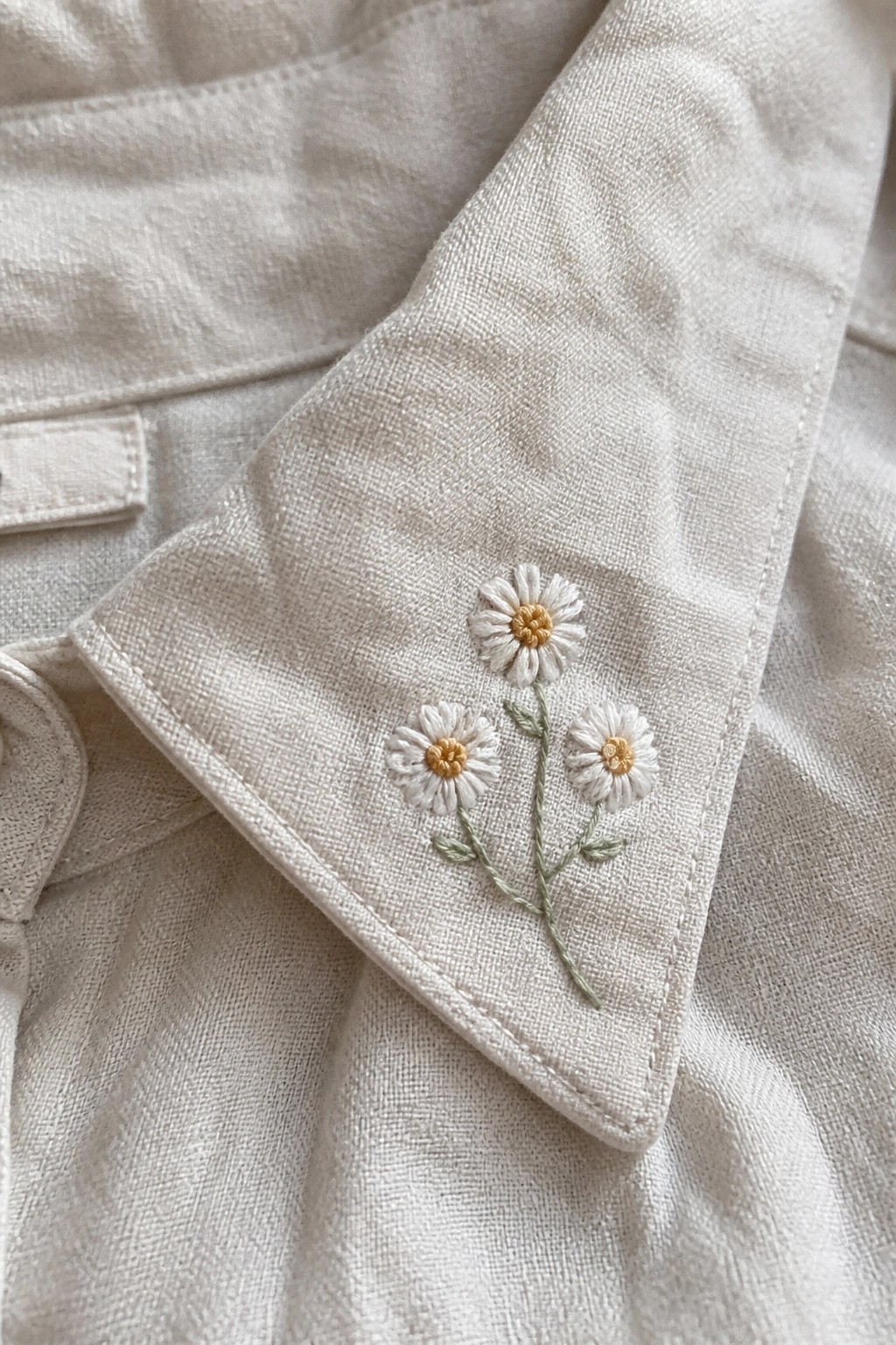 Three embroidered daisies with white petals, yellow centers, and green stems on off-white fabric near a folded edge.