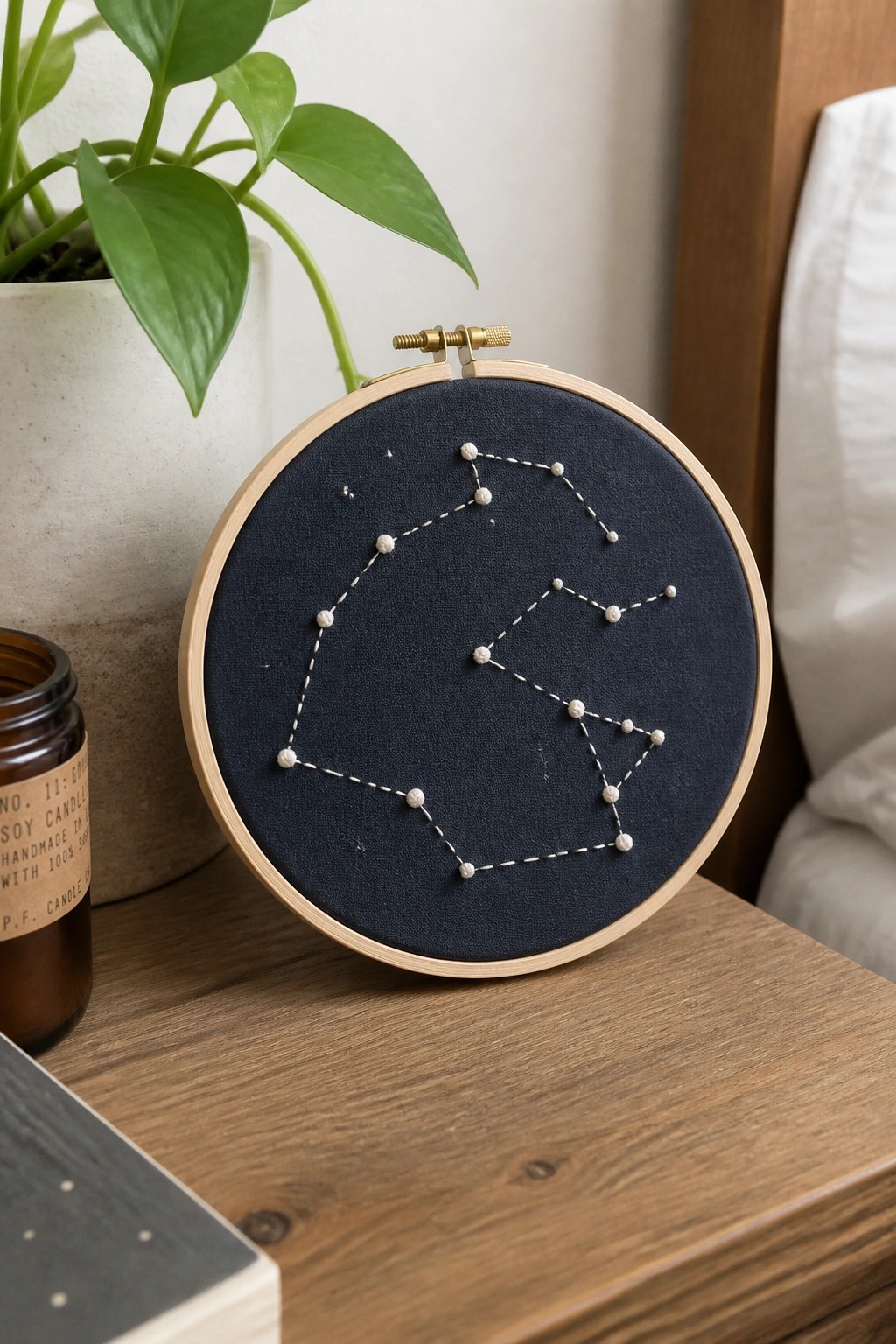 Constellation embroidery in wooden hoop on table with plant, candle, and book.