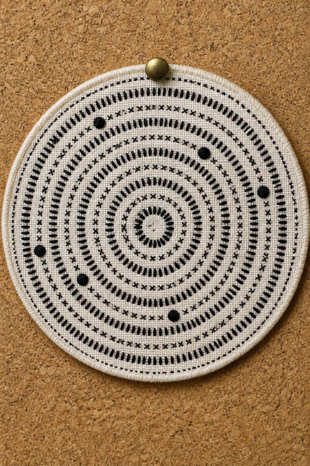 White circular embroidery with black spirals, dashes, and dots pinned to corkboard.
