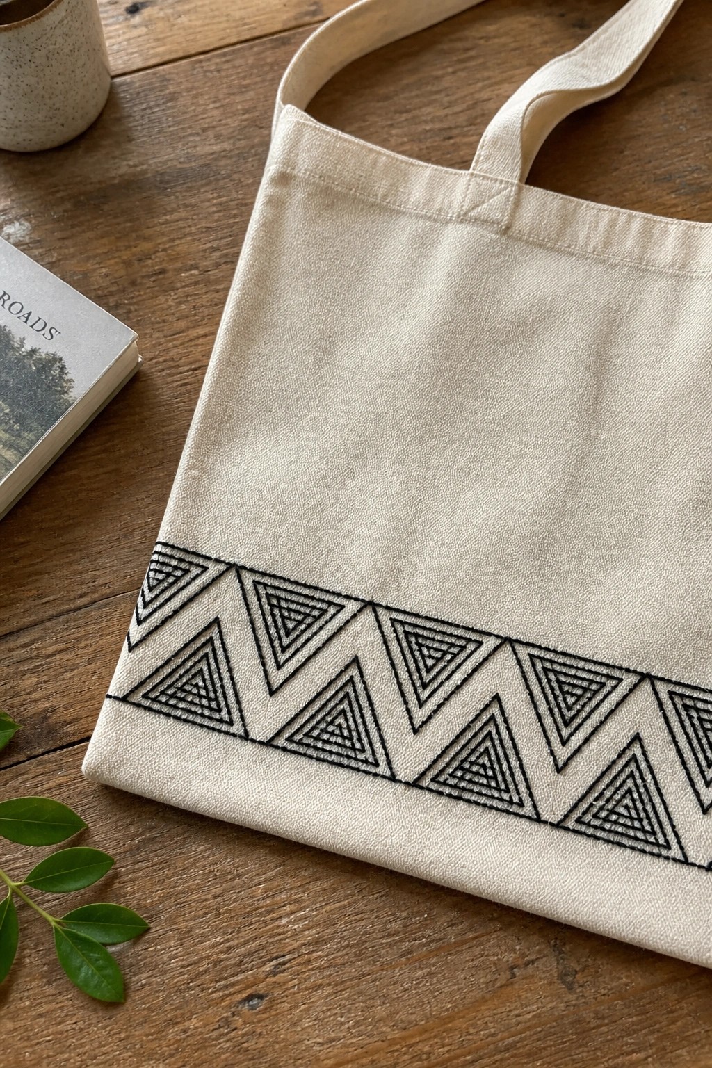 Beige canvas tote bag with black triangular embroidery on wooden table with book