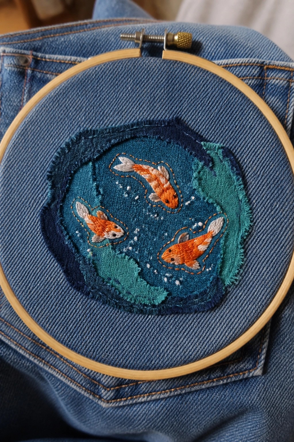 Three orange koi fish embroidered on blue denim patch in wooden hoop