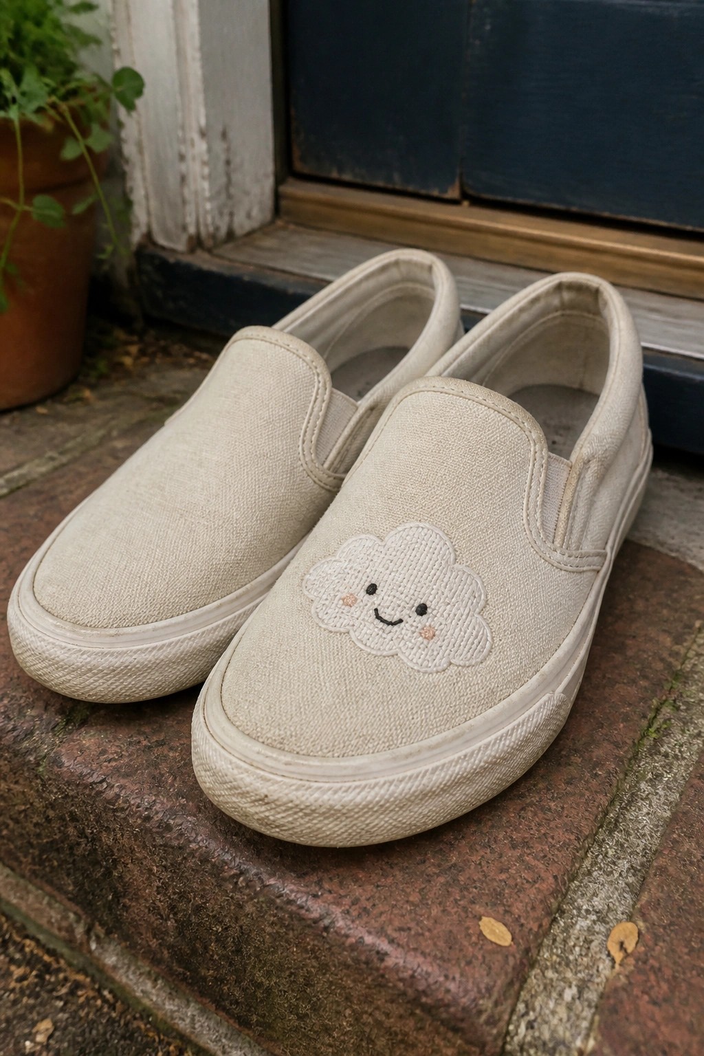 Beige canvas slip-on sneakers with smiling cloud embroidery on brick steps by blue door
