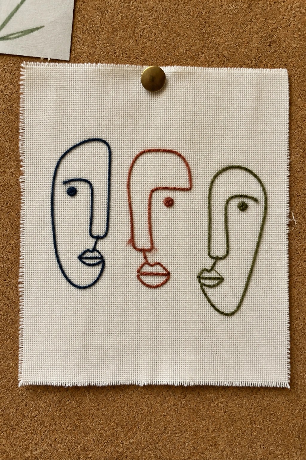 Three abstract faces embroidered in blue, red, and green on pinned fabric
