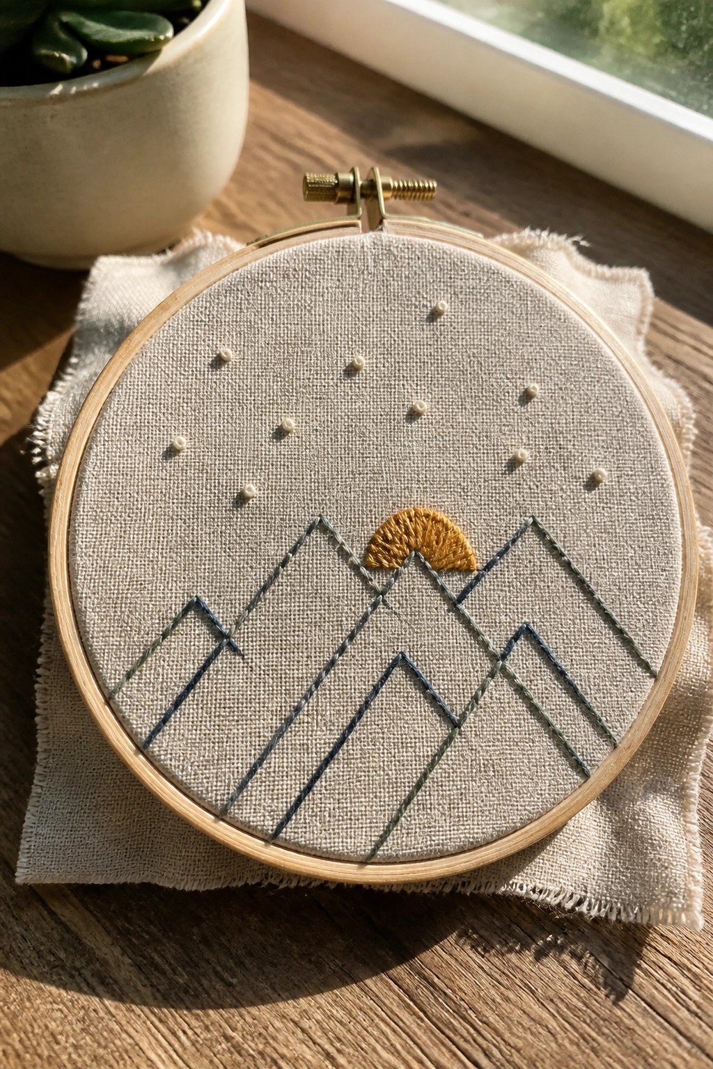 Embroidery hoop with beige fabric showing blue-green mountains, a yellow sun, and scattered white dots.
