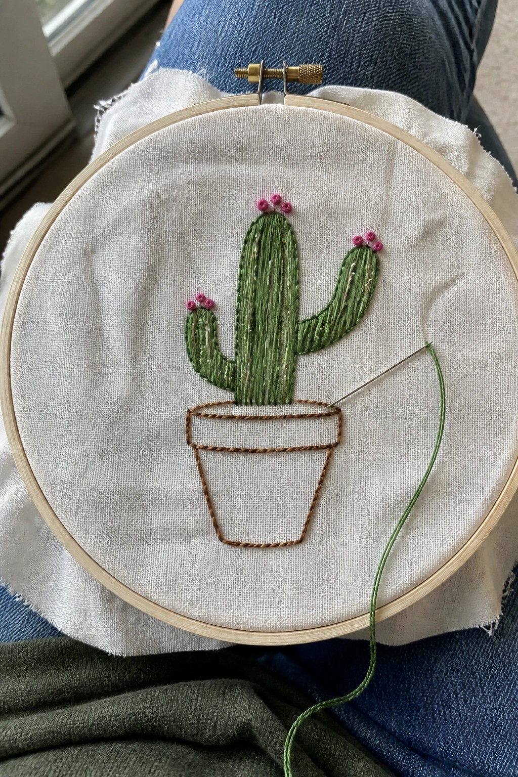 Wooden hoop with cactus embroidery, pink flowers, brown pot, needle and green thread on jeans