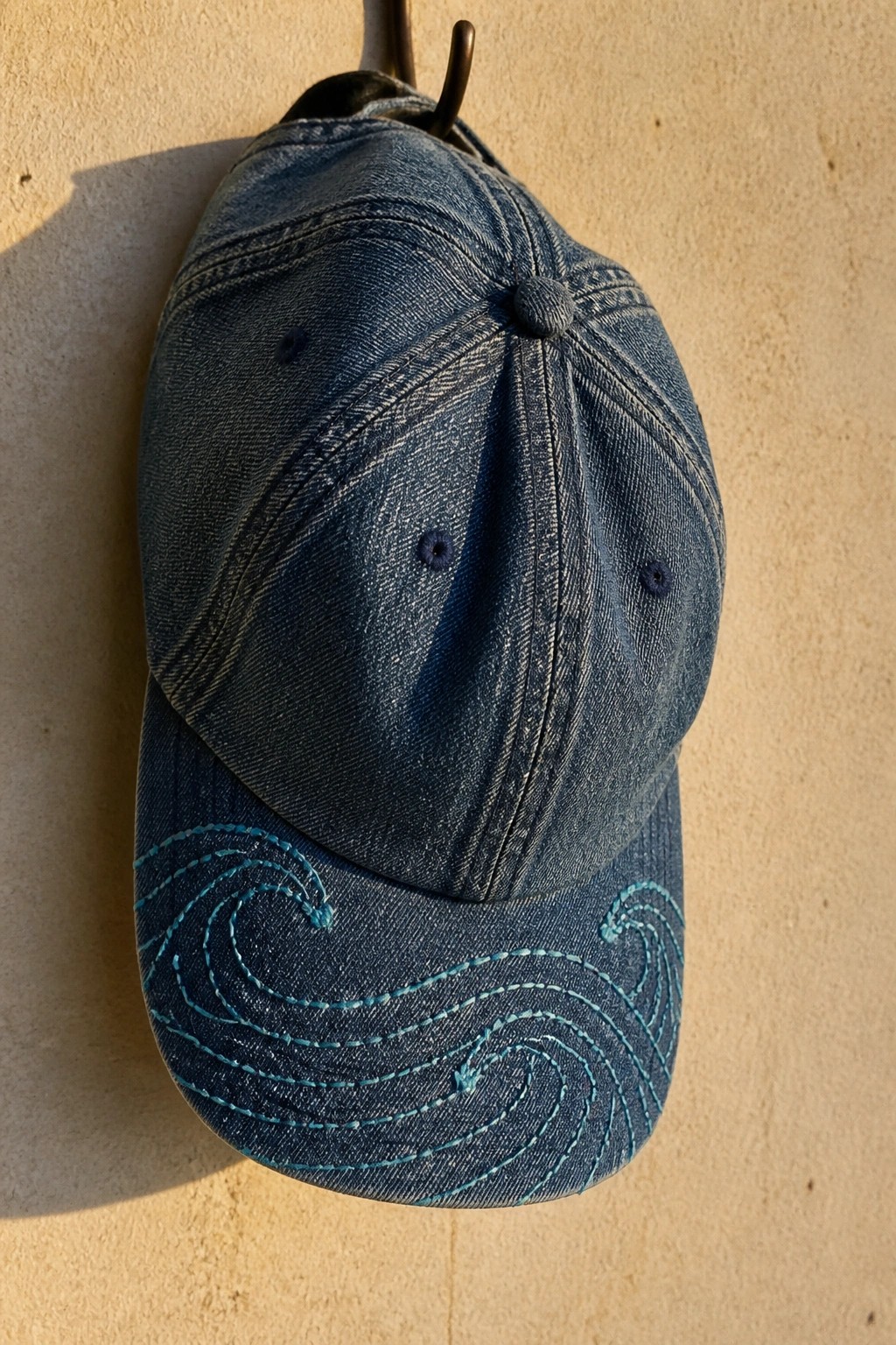 Blue denim baseball cap with turquoise wave embroidery hanging on metal hook
