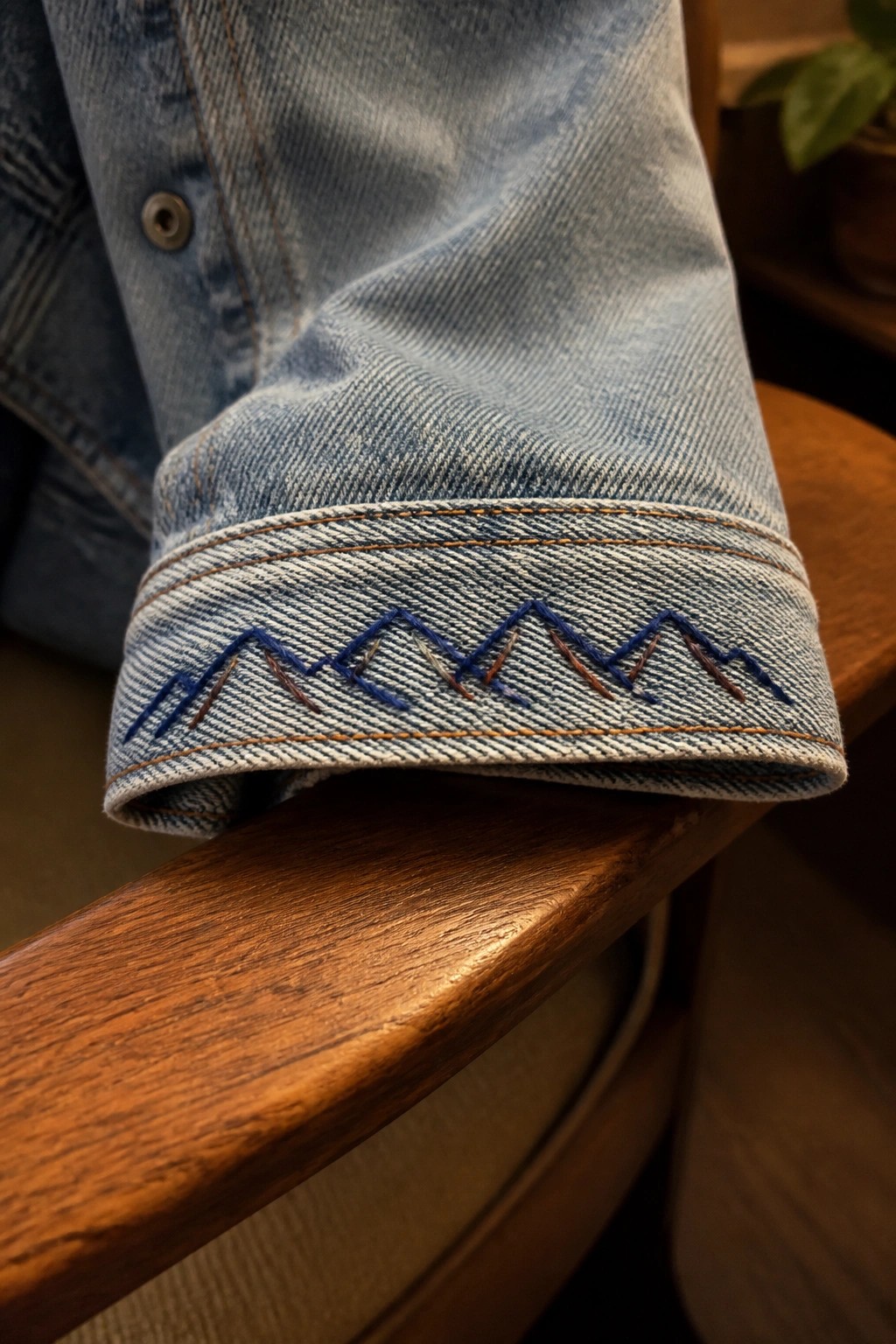Faded blue denim jacket cuff with blue-brown zigzag embroidery on wooden armrest.